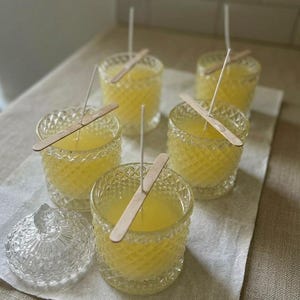 May include: A collection of handmade candles in diamond-cut glass jars. Each candle has a yellow wax fill, a wooden wick holder, and a white wick. A clear glass jar with a lid is also present. The candles are arranged on a neutral-colored cloth.