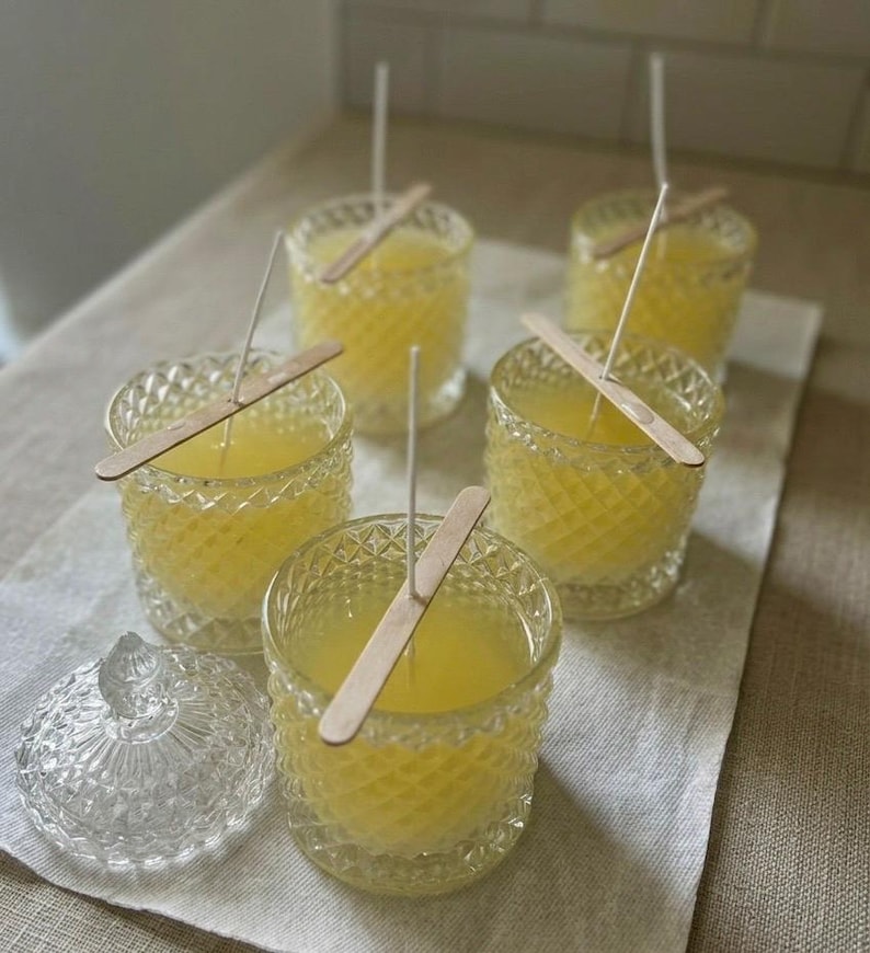 May include: A collection of handmade candles in diamond-cut glass jars. Each candle has a yellow wax fill, a wooden wick holder, and a white wick. A clear glass jar with a lid is also present. The candles are arranged on a neutral-colored cloth.