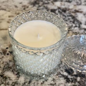 May include: A clear glass candle holder with a diamond pattern, filled with a creamy white candle. The lid is off to the side. The candle has a single wick. The candle holder is on a speckled surface.