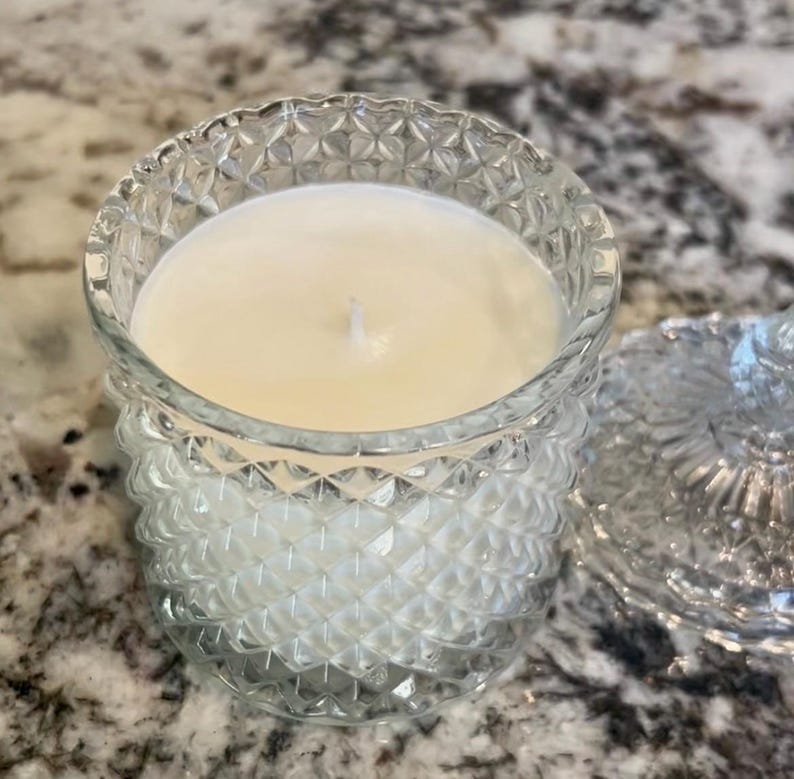 May include: A clear glass candle holder with a diamond pattern, filled with a creamy white candle. The lid is off to the side. The candle has a single wick. The candle holder is on a speckled surface.