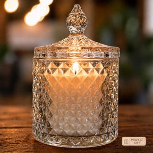 May include: A lit, clear glass candle with a diamond pattern and a lid. The candle is in a jar with a diamond pattern. The flame is lit, and the candle is on a wooden surface. The candle is a perfect gift.