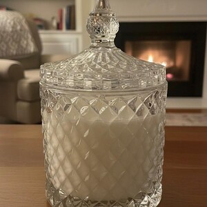 May include: A clear glass candle jar with a diamond pattern and a decorative lid. The jar is filled with a white candle. The background includes a fireplace and a wooden table.