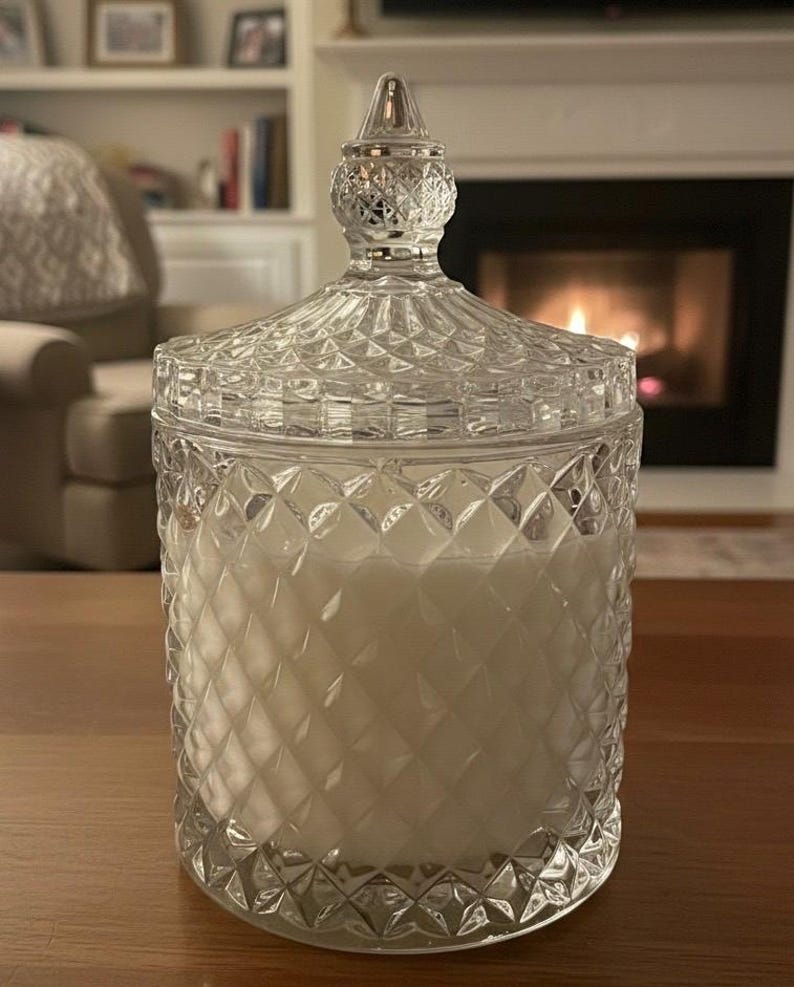 May include: A clear glass candle jar with a diamond pattern and a decorative lid. The jar is filled with a white candle. The background includes a fireplace and a wooden table.