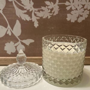 May include: A clear glass candle jar with a diamond pattern, lid off to the side. The candle inside is white. The lid has a decorative finial. The background is a brown wooden board with white floral designs.
