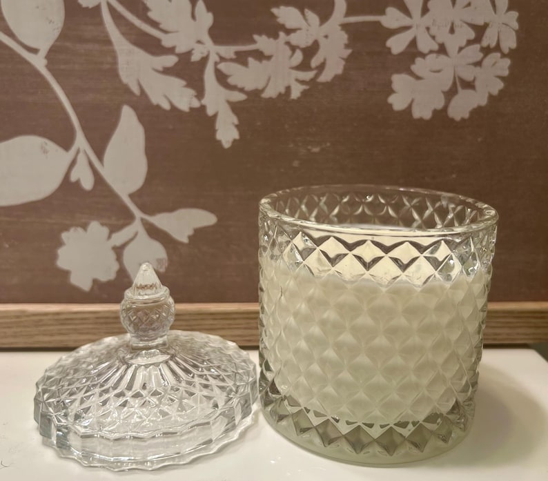 May include: A clear glass candle jar with a diamond pattern, lid off to the side. The candle inside is white. The lid has a decorative finial. The background is a brown wooden board with white floral designs.