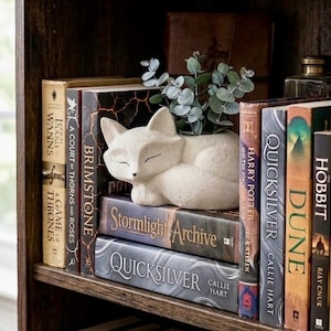 May include: A white fox-shaped planter holding greenery, resting on a stack of books. The books have titles like "Brimstone", "Quicksilver", and "Dune". The bookshelf is made of dark wood, creating a cozy, inviting atmosphere.