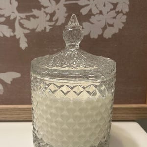 May include: A clear glass candle jar with a diamond pattern and a decorative lid. The jar is filled with a white candle. The candle jar is a decorative home decor item.