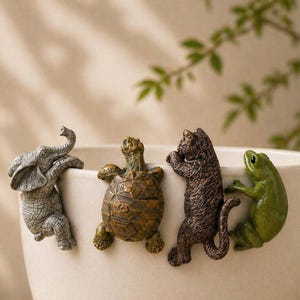 May include: A set of four ceramic animal figurines: an elephant, a turtle, a cat, and a frog. The elephant is gray, the turtle is brown, the cat is dark brown, and the frog is green. Each figurine is designed to hang on the edge of a cup or bowl.