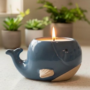 May include: A blue whale-shaped ceramic candle holder with a lit candle. The whale has a light beige accent on its belly and tail. The candle flame is bright against the blue ceramic. The background features potted plants.