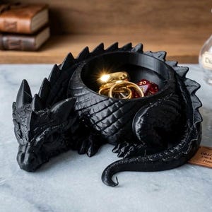 May include: A black dragon-shaped bowl with textured scales, a pointed tail, and horns. The hollow center contains several gold-colored metal rings and a red multi-sided die with white numbers, including a visible number 20.