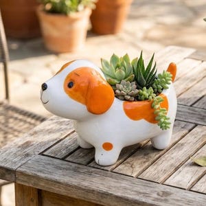 May include: A ceramic dog-shaped planter with a white base and orange spots and ears. The planter is filled with various green succulents. The dog has a smiling face and a small tail. The planter is sitting on a wooden table.
