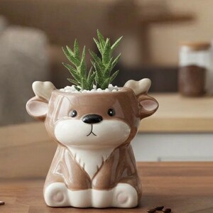 May include: A brown ceramic deer-shaped planter with a white belly and antlers. The planter holds green succulents and white pebbles. The deer has a black nose and a smiling face. The planter is sitting on a wooden surface.