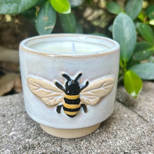 May include: A white ceramic candle jar with a bee design. The bee is brown, black, and yellow. The candle is white and has a wick.