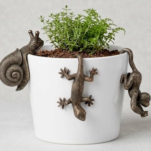 May include: A white ceramic plant pot featuring three bronze-colored metal animal figures attached to the exterior: a snail on the left rim, a lizard climbing the center, and a monkey hanging by its tail from the right rim, containing a green plant.