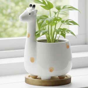 May include: A white ceramic giraffe planter with orange spots, holding a green plant with large leaves. The planter sits on a round wooden base. The giraffe's head forms the handle.