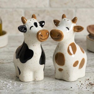 May include: A pair of ceramic cow-shaped salt and pepper shakers. One cow is white with black spots, the other is white with brown spots. Both have brown ears and noses, and smiling faces. The shakers are on a speckled surface.