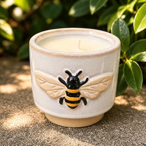 May include: A ceramic candle in a beige pot with a raised bee design. The bee has black and yellow stripes and tan wings. The candle wax is a creamy color, and the pot has a textured finish.