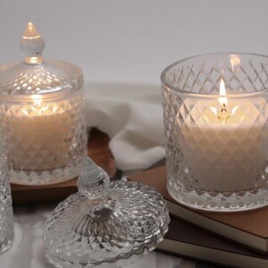 May include: Clear glass candle holders with diamond-patterned designs. One candle is lit, showing a warm glow. The lid of one holder is off, revealing the candle inside. The candles sit on a wooden surface and a stack of books.