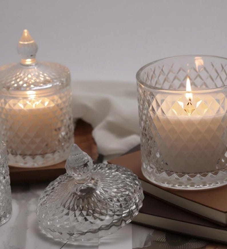May include: Clear glass candle holders with diamond-patterned designs. One candle is lit, showing a warm glow. The lid of one holder is off, revealing the candle inside. The candles sit on a wooden surface and a stack of books.