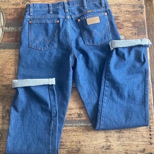 Może przedstawiać: Niebieskie dżinsy z denimu o klasycznym kroju. Posiadają miedziane nity, sk&oacute;rzaną naszywkę z logo "Wrangler" i charakterystyczne przeszycia tylnych kieszeni. Mankiety są podwinięte, ukazując jaśniejszy szary materiał.