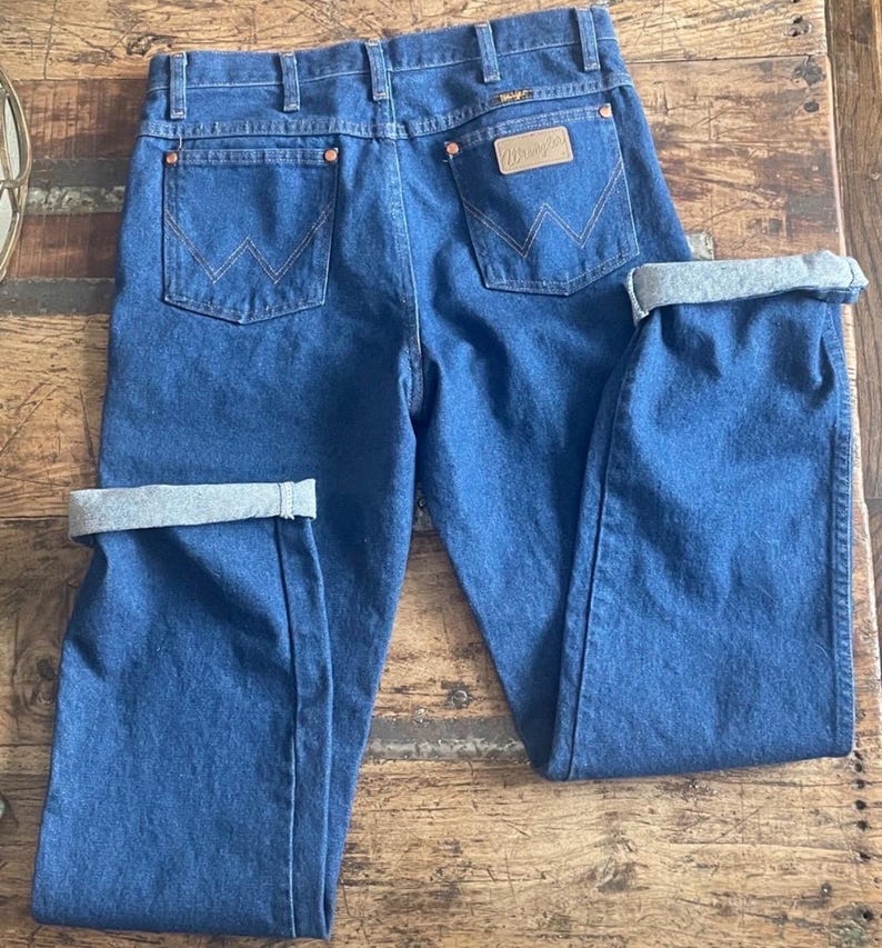 Może przedstawiać: Niebieskie dżinsy z denimu o klasycznym kroju. Posiadają miedziane nity, sk&oacute;rzaną naszywkę z logo "Wrangler" i charakterystyczne przeszycia tylnych kieszeni. Mankiety są podwinięte, ukazując jaśniejszy szary materiał.