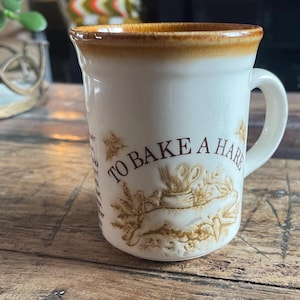 May include: Cream-colored ceramic mug with a brown rim and handle. The mug features a brown illustration of a rabbit, wheat, and the text "TO BAKE A HARE". The mug is on a wooden surface.