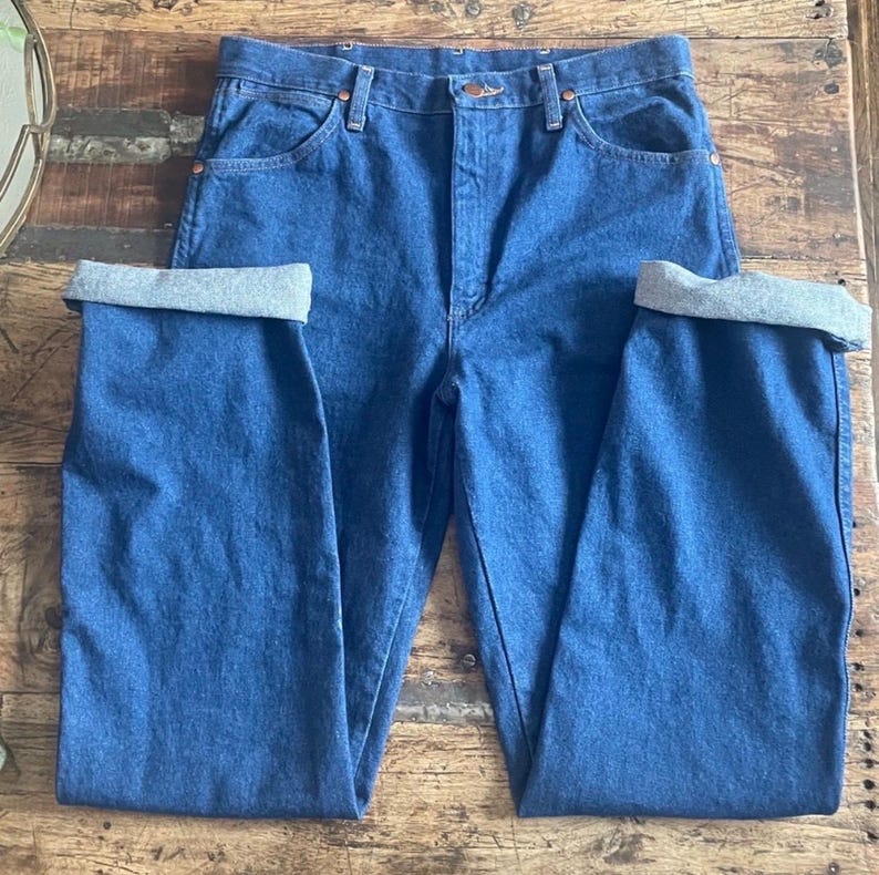 Może przedstawiać: Niebieskie dżinsy z denimu z podwiniętymi mankietami. Dżinsy mają klasyczny pięciokieszeniowy design z miedzianymi nitami i zapięciem na guzik. Denim jest w średnim odcieniu, a mankiety odsłaniają szare wnętrze.