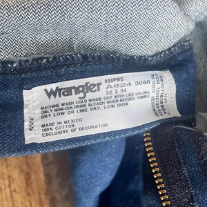 Może przedstawiać: Zbliżenie etykiety dżins&oacute;w Wrangler. Biała etykieta zawiera czarny tekst z instrukcjami prania, rozmiarem 32 x 34 i informacjami o materiale. Dżinsy są wykonane z ciemnoniebieskiego denimu z widocznym zamkiem błyskawicznym.
