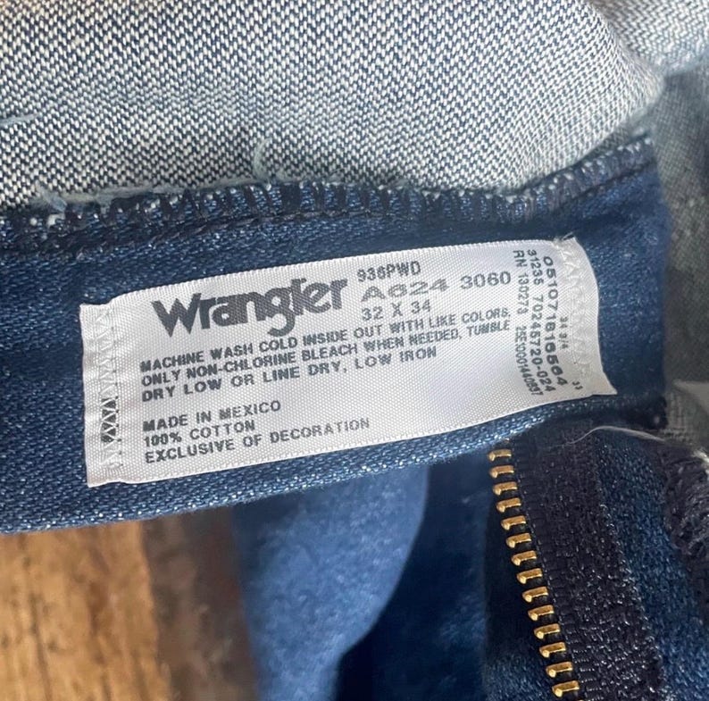 Może przedstawiać: Zbliżenie etykiety dżins&oacute;w Wrangler. Biała etykieta zawiera czarny tekst z instrukcjami prania, rozmiarem 32 x 34 i informacjami o materiale. Dżinsy są wykonane z ciemnoniebieskiego denimu z widocznym zamkiem błyskawicznym.