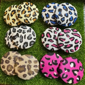May include: A collection of eight round fabric buttons with animal print patterns. The buttons feature leopard spots in various colors, including yellow, brown, black, blue, pink, and gray. The buttons are arranged on a green surface.