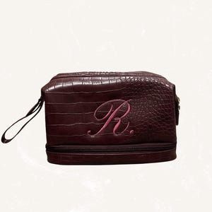 May include: A burgundy leather travel bag with a crocodile texture. The bag features a pink embroidered letter "R" on the front. It has a zipper closure and a small handle on the side. The bag is designed for toiletries or personal items.