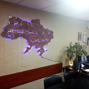May include: A wooden map of Ukraine with purple LED lighting outlining the borders and major cities. The map is mounted on a beige wall, with a dark wooden trim below. The map is connected to a power source.