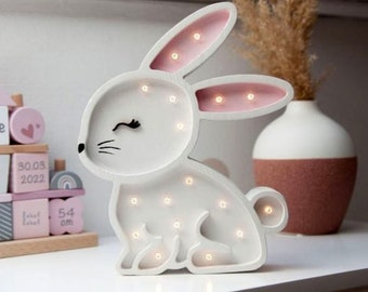 Könnte beinhalten: Weißes, hölzernes Nachtlicht in Form eines Hasen mit rosa Ohren und weißen Punkten. Der Hase hat ein schwarzes Auge und Wimpern. Das Licht hat kleine weiße LED-Leuchten um den Körper und die Ohren.