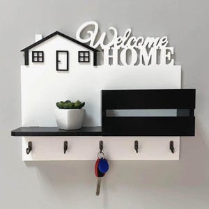 May include: A white and black wooden key holder with a house-shaped design and the words "Welcome HOME". It has a shelf, a small potted plant, a mail slot, and hooks for keys. The keys hanging from the hooks have blue and red keychains.