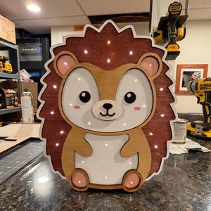 Pode incluir: Uma luz noturna de madeira em forma de ouriço com um design castanho, castanho claro e branco. O ouriço tem uma borda externa espinhosa castanha escura, um rosto castanho claro e uma barriga branca. Pequenas luzes estão embutidas.