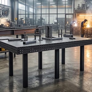 Könnte beinhalten: Ein großer, dunkelgrauer Metall-Schweißtisch mit perforierter Oberfläche und schwarzen Beinen. Auf dem Tisch sind mehrere Metallteile angeordnet. Schweißhelme stehen auf einem Regal im Hintergrund. Die Szene spielt in einer Werkstatt.