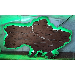 Puede incluir: Un mapa de madera de Ucrania con nombres de ciudades grabados. El mapa está delineado con una luz LED verde brillante, creando un efecto brillante. Está montado en una pared de ladrillos blancos.