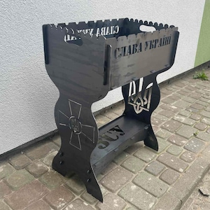 May include: A black metal barbecue grill with cut-out designs and the Ukrainian coat of arms. The grill features the words "Glory to Ukraine" and other text. It has a decorative base and a rectangular firebox.