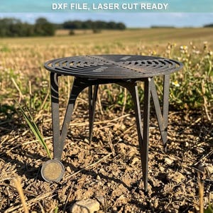 May include: A dark gray metal circular stand with a laser-cut concentric ring pattern on the top surface and four pointed legs. The text DXF FILE, LASER CUT READY is visible at the top. A 2 Euro coin is placed next to the stand for scale.