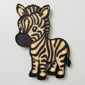 May include: A wooden zebra night light with black and white stripes and a black outline. The zebra has a cute face with black eyes and a black nose. The night light has small, round, yellow lights that illuminate the zebra's body.