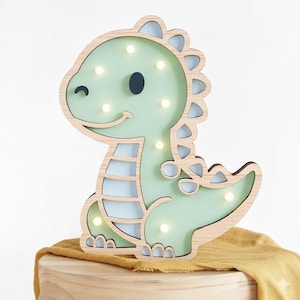 Może przedstawiać: Drewniana lampka nocna w kształcie dinozaura z jasnozielonym korpusem i jasnoniebieskim wnętrzem. Dinozaur ma uśmiechniętą twarz i rząd małych światełek. Światło stoi na drewnianej powierzchni.