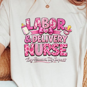 Puede incluir: Camiseta color crema con un gráfico rosa que dice "LABOR & DELIVERY NURSE" con una ilustración de biberón y chupete. El texto "Tiny Humans, Big Impact" está debajo. El diseño presenta un patrón de corazón.