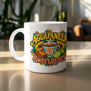 Könnte beinhalten: Ein weißer Keramikbecher mit einer farbenfrohen grafischen Illustration einer dampfenden Tasse Aguapanela, zwei braunen Zuckerwürfeln, einer Limettenscheibe und Sonnenblumen. Der Text AGUAPANELA und SWEET RESET ist in fetten, stilisierten Buchstaben gedruckt.