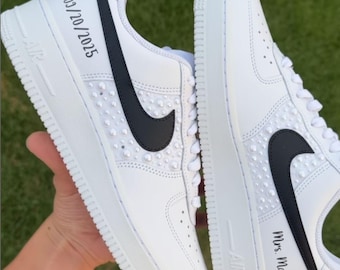 May include: White sneakers with black swooshes and embellishments. One shoe has "03/20/2025" printed on the side, the other has "Mrs. Martins". The shoes are on a green background.