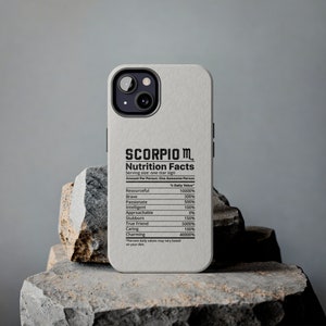 May include: A light gray phone case with a black camera and a printed "Scorpio Nutrition Facts" label. The label lists personality traits like "Resourceful" and "Charming" with corresponding percentage values. The phone case is displayed on a stone surface.