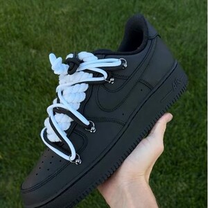 K&ouml;nnte beinhalten: Schwarze AF1 "Black Panda" Sneaker mit wei&szlig;en und schwarzen Seilschn&uuml;rsenkeln. Die Schuhe werden vor einem gr&uuml;nen Gras Hintergrund gehalten. Die Sneaker haben eine schwarze Sohle und ein schwarzes Obermaterial.