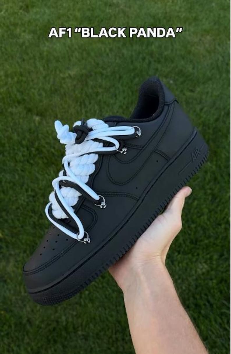 K&ouml;nnte beinhalten: Schwarze AF1 "Black Panda" Sneaker mit wei&szlig;en und schwarzen Seilschn&uuml;rsenkeln. Die Schuhe werden vor einem gr&uuml;nen Gras Hintergrund gehalten. Die Sneaker haben eine schwarze Sohle und ein schwarzes Obermaterial.