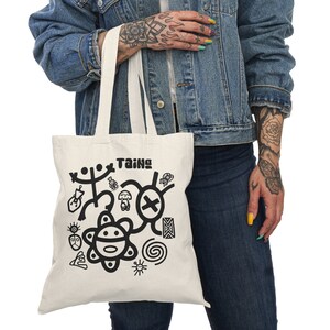 May include: A white tote bag with black handles and a black design of Taíno symbols, including a sun, a spiral, and a cross. The text "Taíno" is printed in black at the top of the bag.