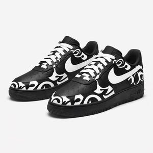May include: Pair of black and white sneakers. The shoes feature a black base with white decorative patterns and a white Nike swoosh. White laces complete the design. The sneakers are on a white background.