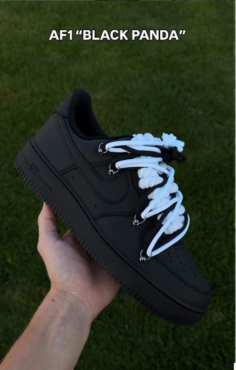 K&ouml;nnte beinhalten: Schwarze Nike Air Force 1 "Black Panda" Sneaker mit wei&szlig;en Schn&uuml;rsenkeln. Die Schuhe sind ganz schwarz mit einem wei&szlig;-schwarzen Schn&uuml;rsenkeldesign. Der Text "AF1 "BLACK PANDA"" befindet sich oben im Bild.